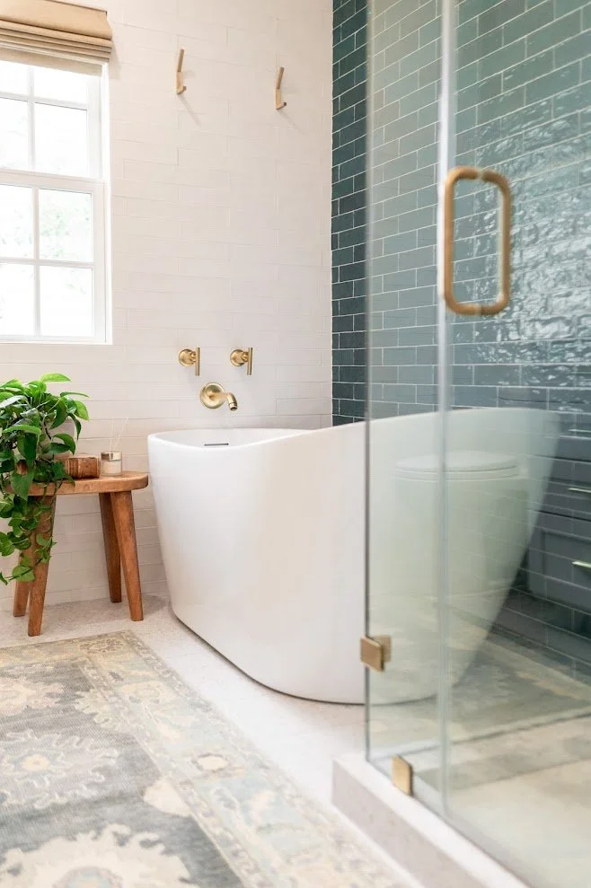 A modern bathroom with a standalone bathtub, glass shower enclosure with a brass handle, a wooden side table, a green plant, and a patterned rug. The room has light-colored walls, a window with shades, and gold fixtures.