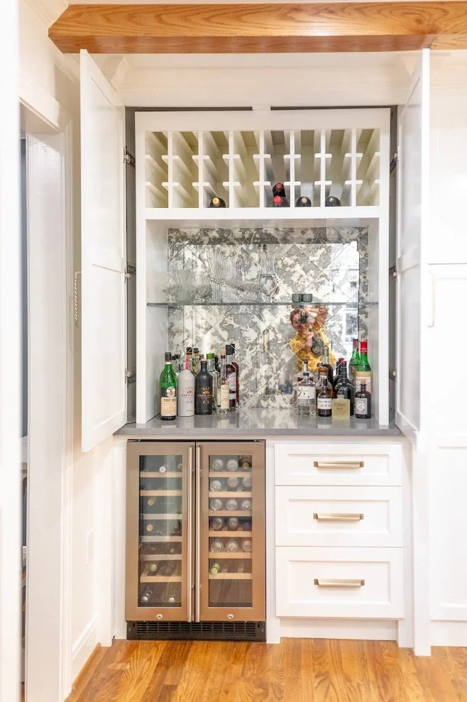 Built-in home bar cabinet with open wine rack at the top, glass shelves with liquor bottles, and a wine cooler at the bottom, set against a marble wall with a floral decoration.