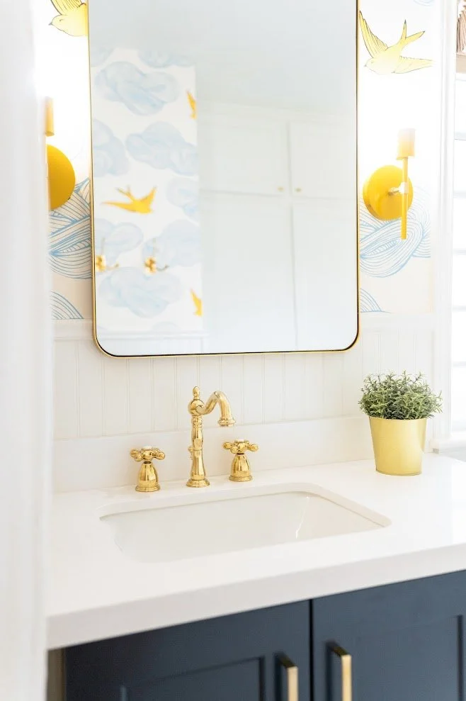 Bathroom vanity with gold fixtures, white countertop, mirror, yellow wall sconces, decorative wall art, and a potted plant.