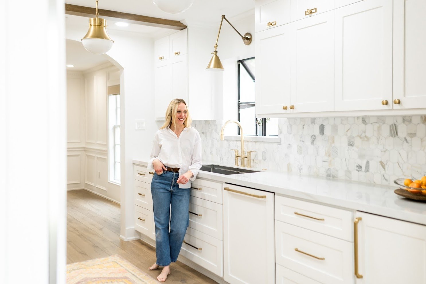 Woman standing in a bright, modern kitchen with white cabinets, gold hardware, marble backsplash, black sink, and gold faucet, smiling and barefoot.