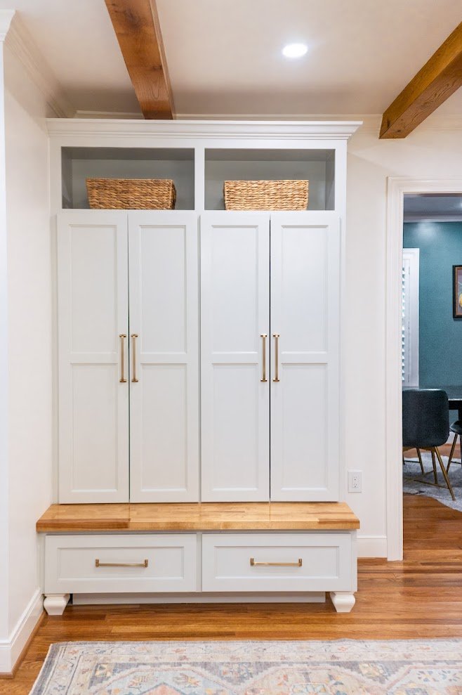 A white built-in cabinet with multiple doors, gold handles, two woven baskets on top, and a wooden bench at the bottom, situated in a room with hardwood flooring and a patterned rug.