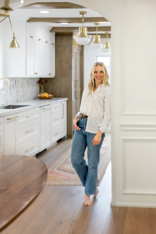 A woman standing in a modern kitchen with white cabinets, gold fixtures, and a wooden floor, smiling and looking at the camera.