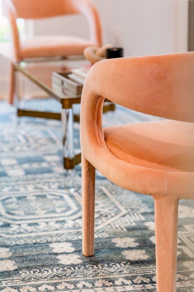 Close-up of a peach-colored upholstered armchair with a curved armrest in a living room with a patterned rug and a coffee table in the background.