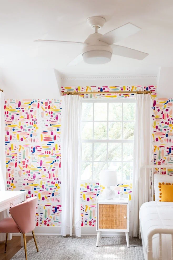 Bright bedroom with colorful abstract wallpaper, white curtains, a white nightstand with a white lamp, and a bed with a yellow pillow.