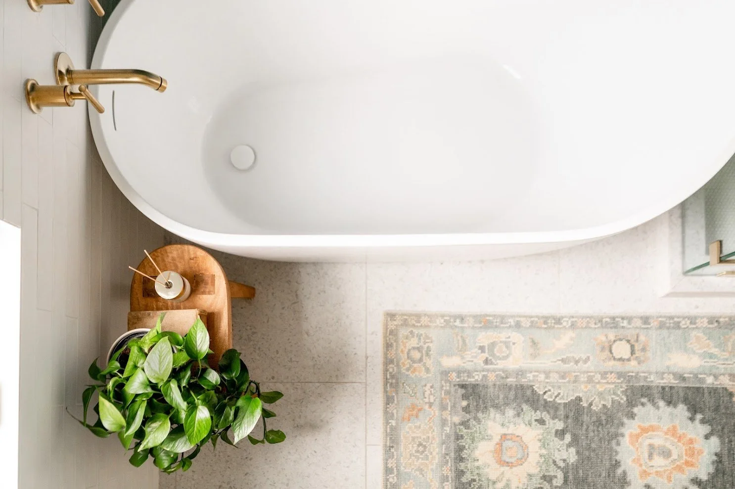 Top-down view of a bathroom corner featuring a white bathtub, a small wooden side table with a potted green plant, a reed diffuser, and a light-colored area rug with floral patterns.
