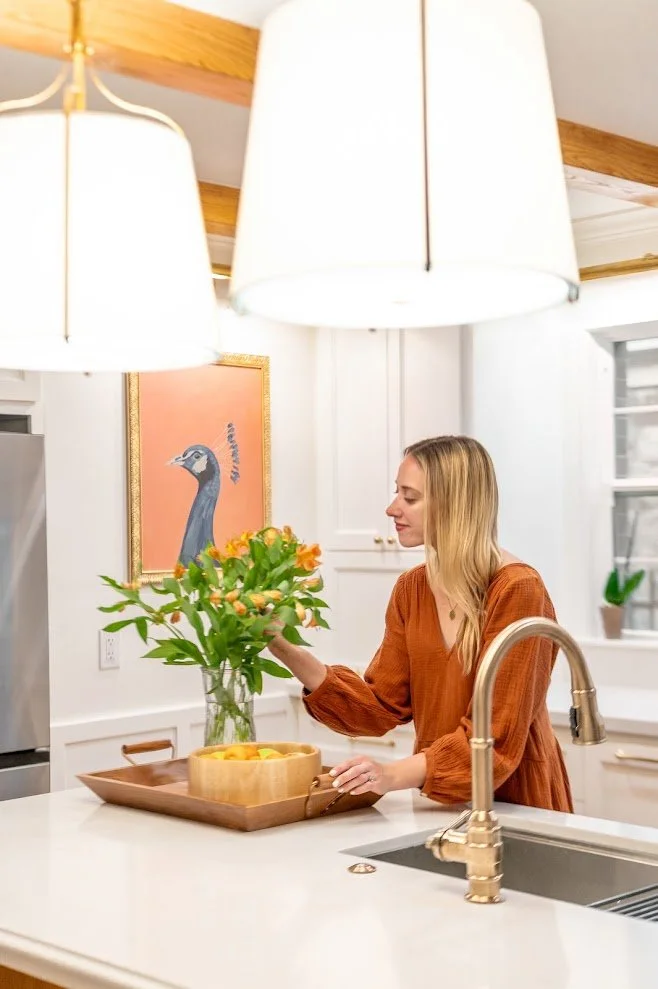 A woman in a burnt orange dress arranging a bouquet of flowers in a glass vase on a kitchen counter with a wooden tray of lemons nearby. There are two large white pendant lights hanging from the ceiling, a framed peacock art on the wall, and a window with a small potted plant on the sill.