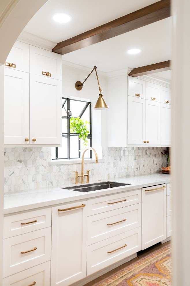 Bright white kitchen with upper and lower cabinets, gold handles, a marble backsplash, a double sink with gold faucet, a window with greenery, and dark wooden ceiling beams.