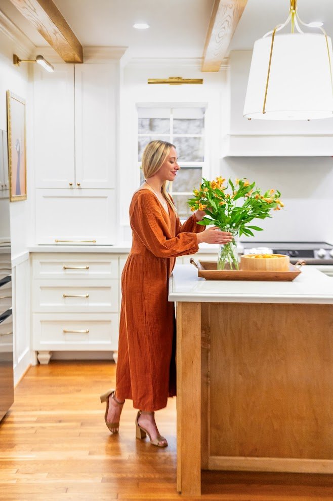 A woman in an orange dress and high heels arranging flowers in a glass vase on a kitchen island.