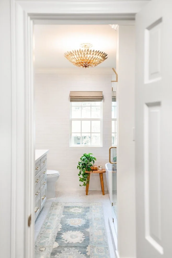 A bright bathroom viewed through a doorway with a white door frame, featuring a window with a beige roller shade, a wooden side table with a green plant, a bathtub with a glass partition, a vanity with gold handles, and a light-colored rug on the flo