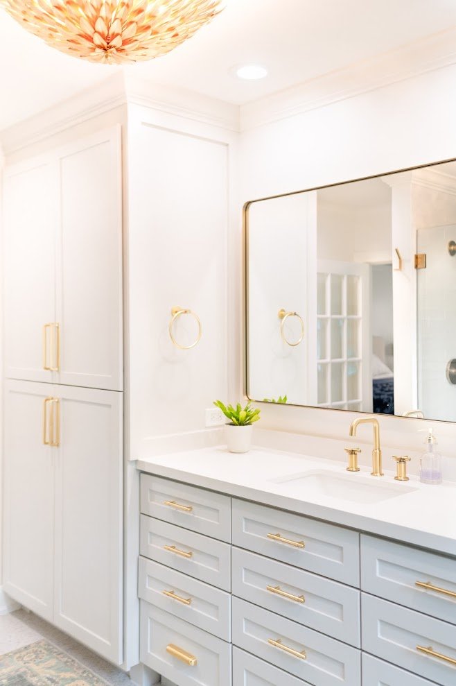 A bright, white bathroom vanity with gold handles and fixtures, a large mirror, a potted green plant, and a soap dispenser, with a built-in cabinet on the side and a decorative ceiling light.