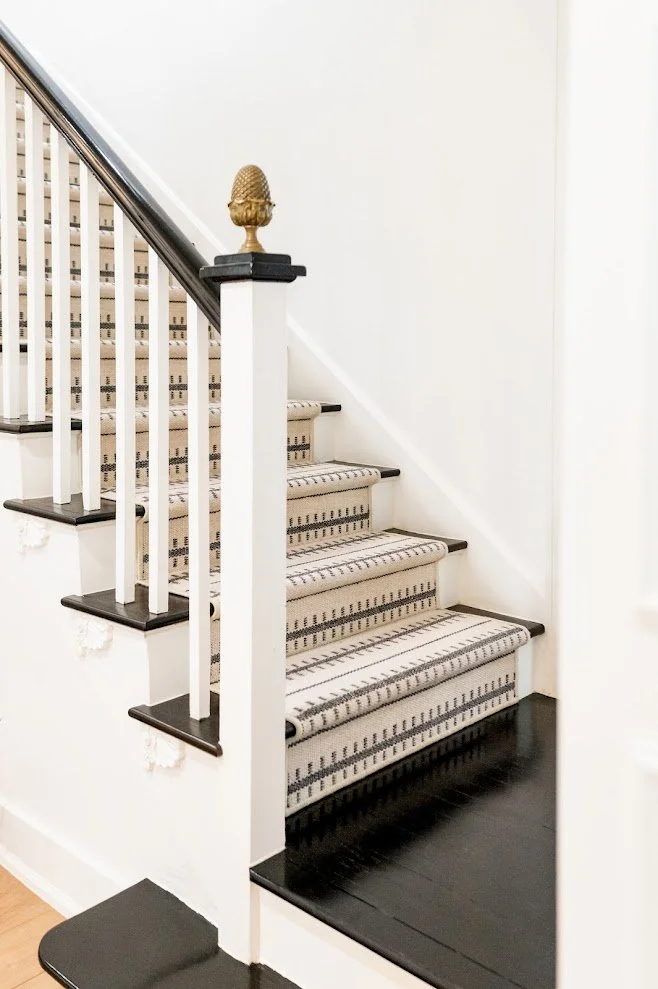Interior staircase with black and beige striped carpet, black treads, white risers, black handrail, decorative gold pineapple finial at the top of the newel post, and black painted floor at the bottom.