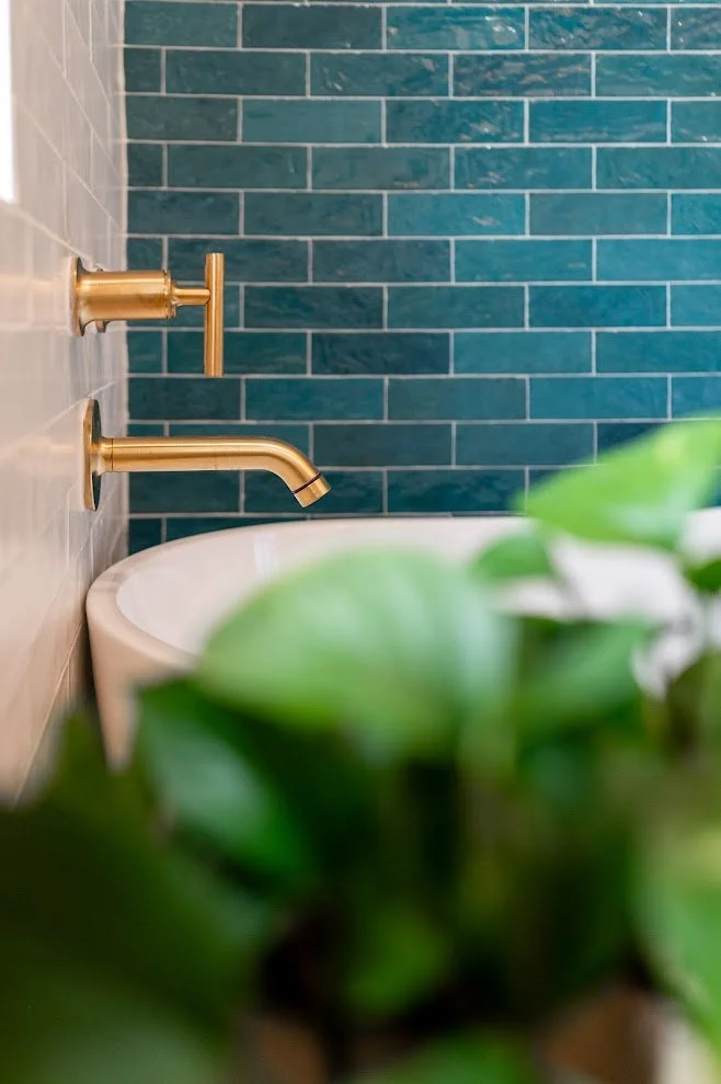 Bathroom with beige and blue tiled walls, gold faucets, and a white bathtub, partially obscured by green plant leaves in the foreground.