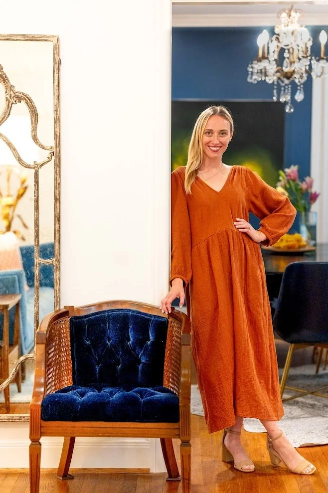 A woman in an orange dress standing in a living room, next to a wooden chair with navy velvet cushion, with a mirror and chandelier in the background.
