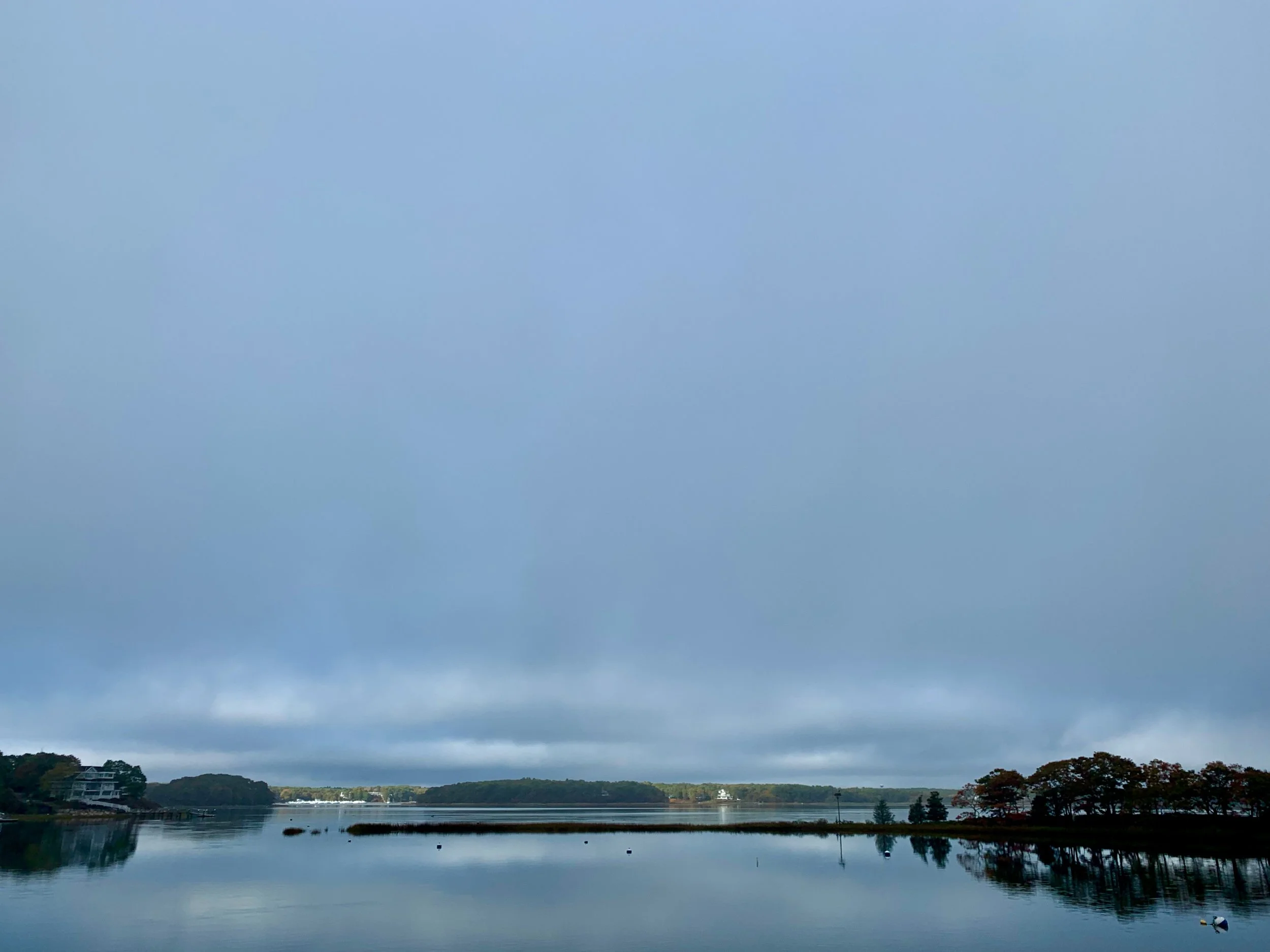 A lake with calm water reflecting cloudy sky, surrounded by trees and a few houses in the distance.