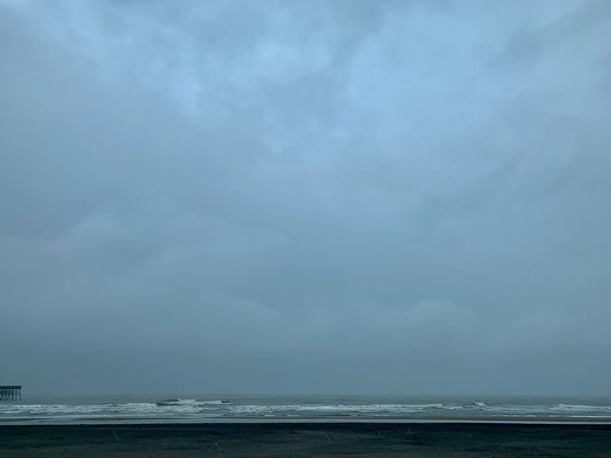 Cloudy sky over a beach with waves and a pier in the distance.