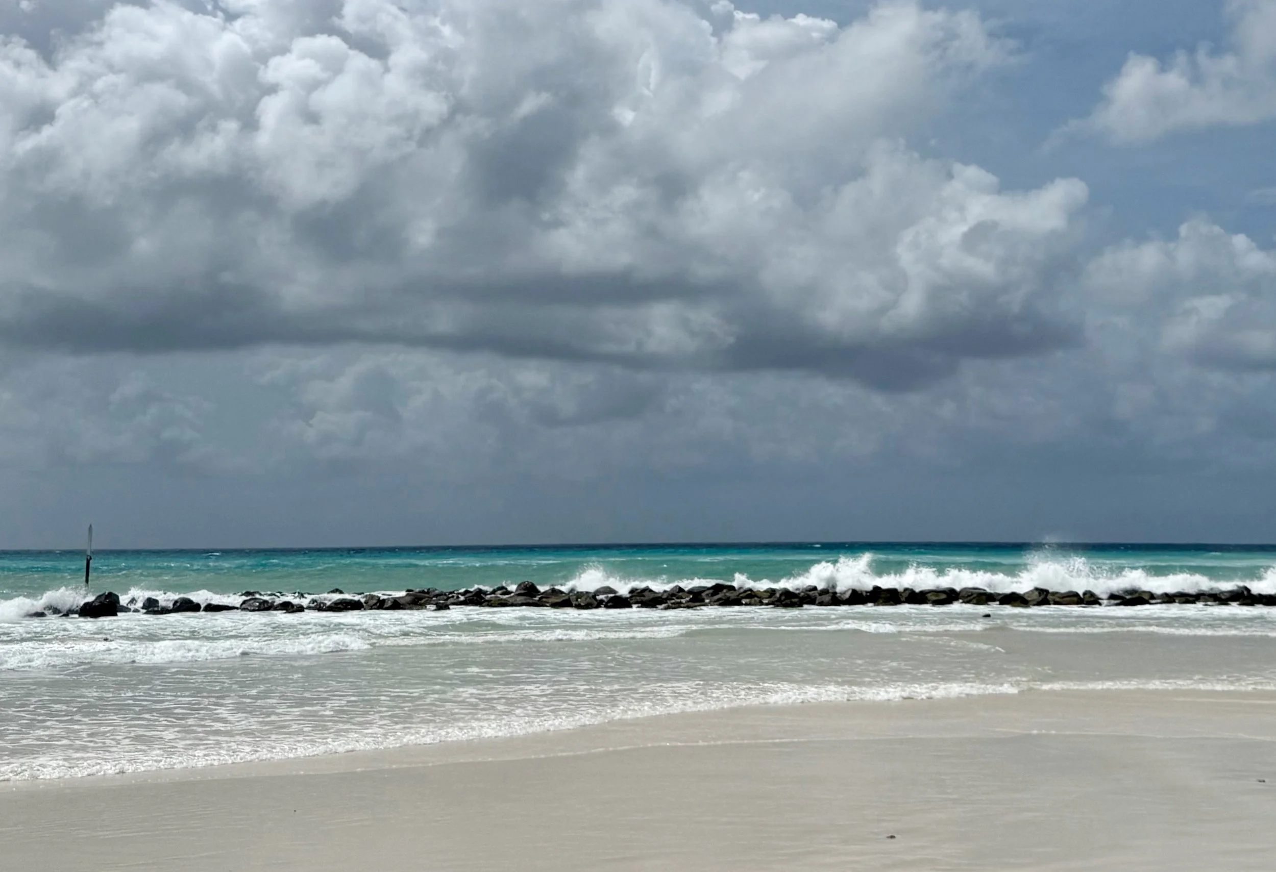 Cloudy sky over a beach with turquoise water, waves hitting a rocky shoreline, and a sailboat far in the distance.