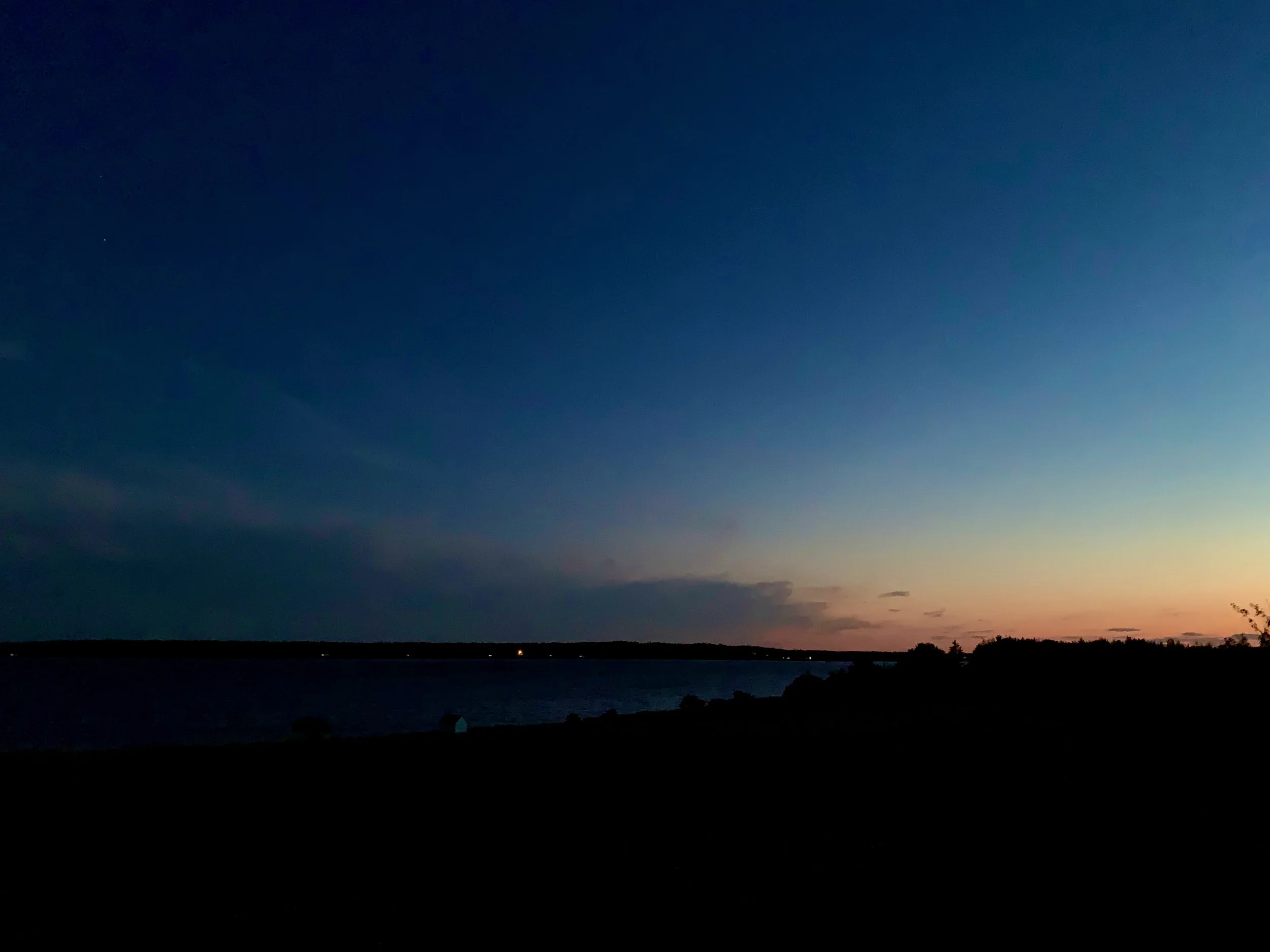 A landscape scene at dusk shows a dark shoreline and water with silhouettes of trees, and a sky transitioning from dark blue to lighter shades with a hint of orange near the horizon.