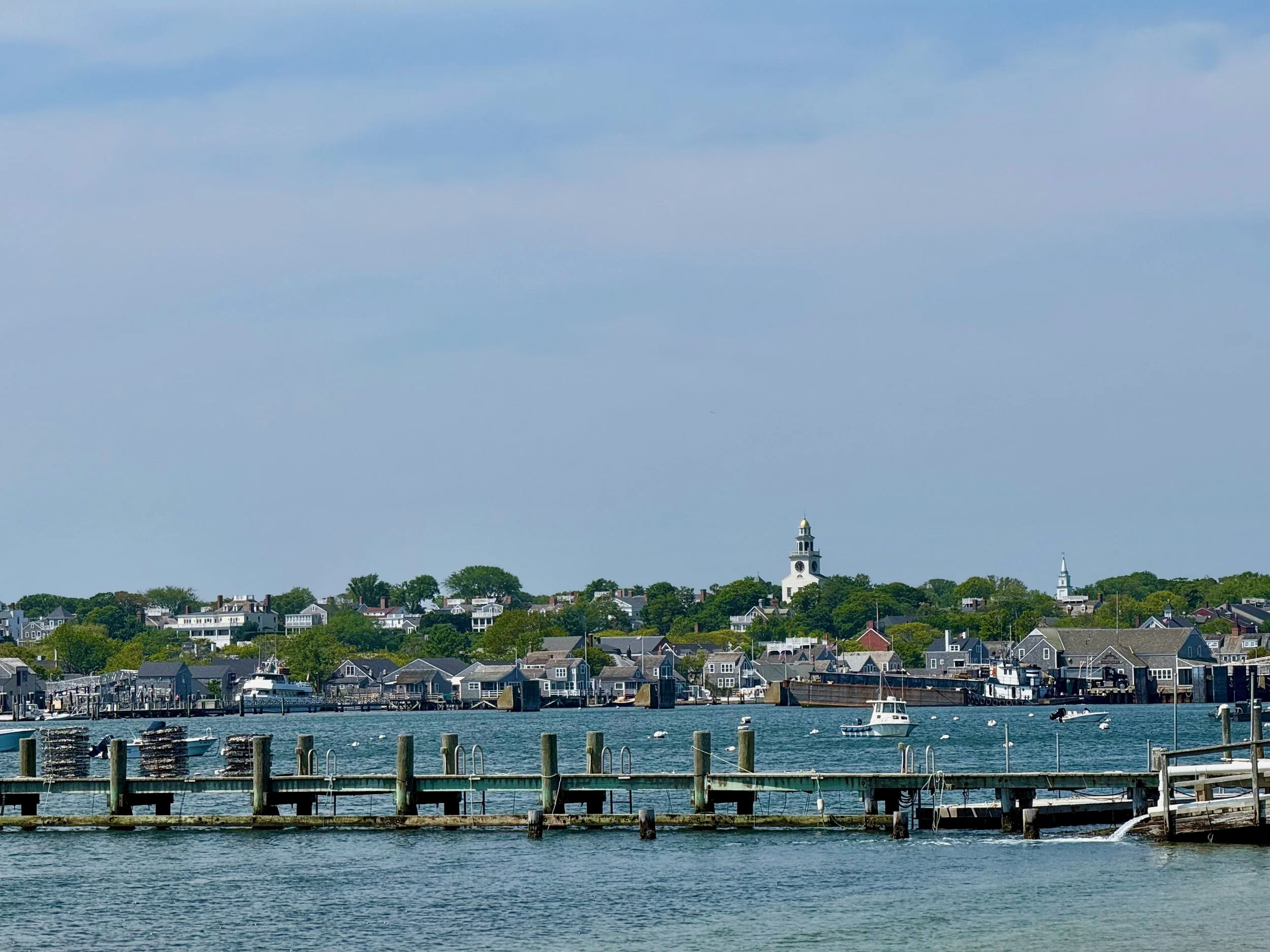 A scenic view of a harbor with boats docked, a shoreline lined with houses and trees, a white lighthouse on a small hill, and a mostly clear sky with some clouds.