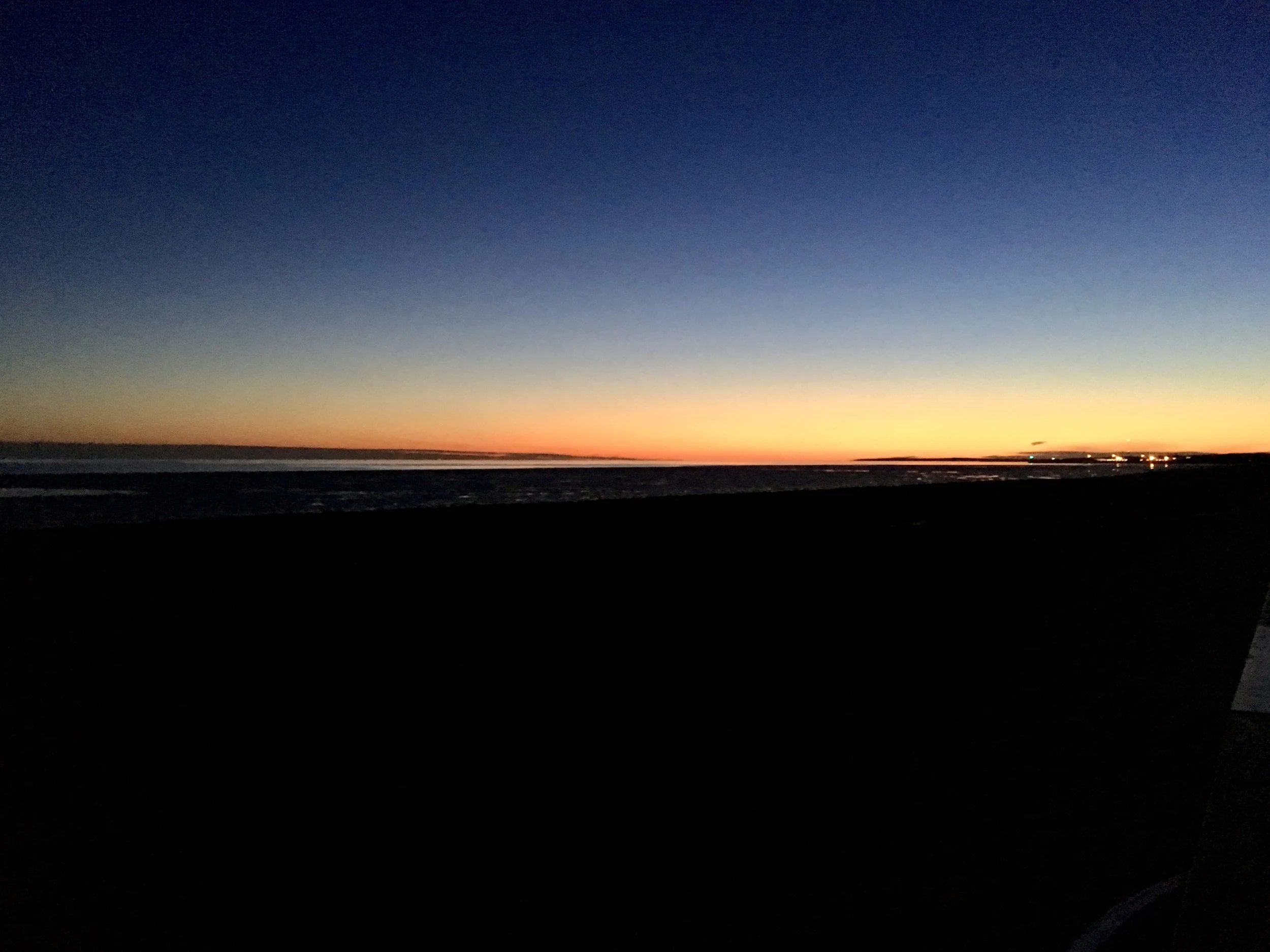 A panoramic view of a sunset over a beach with a large, open sky, colors fading from orange and yellow near the horizon to deep blue higher in the sky.