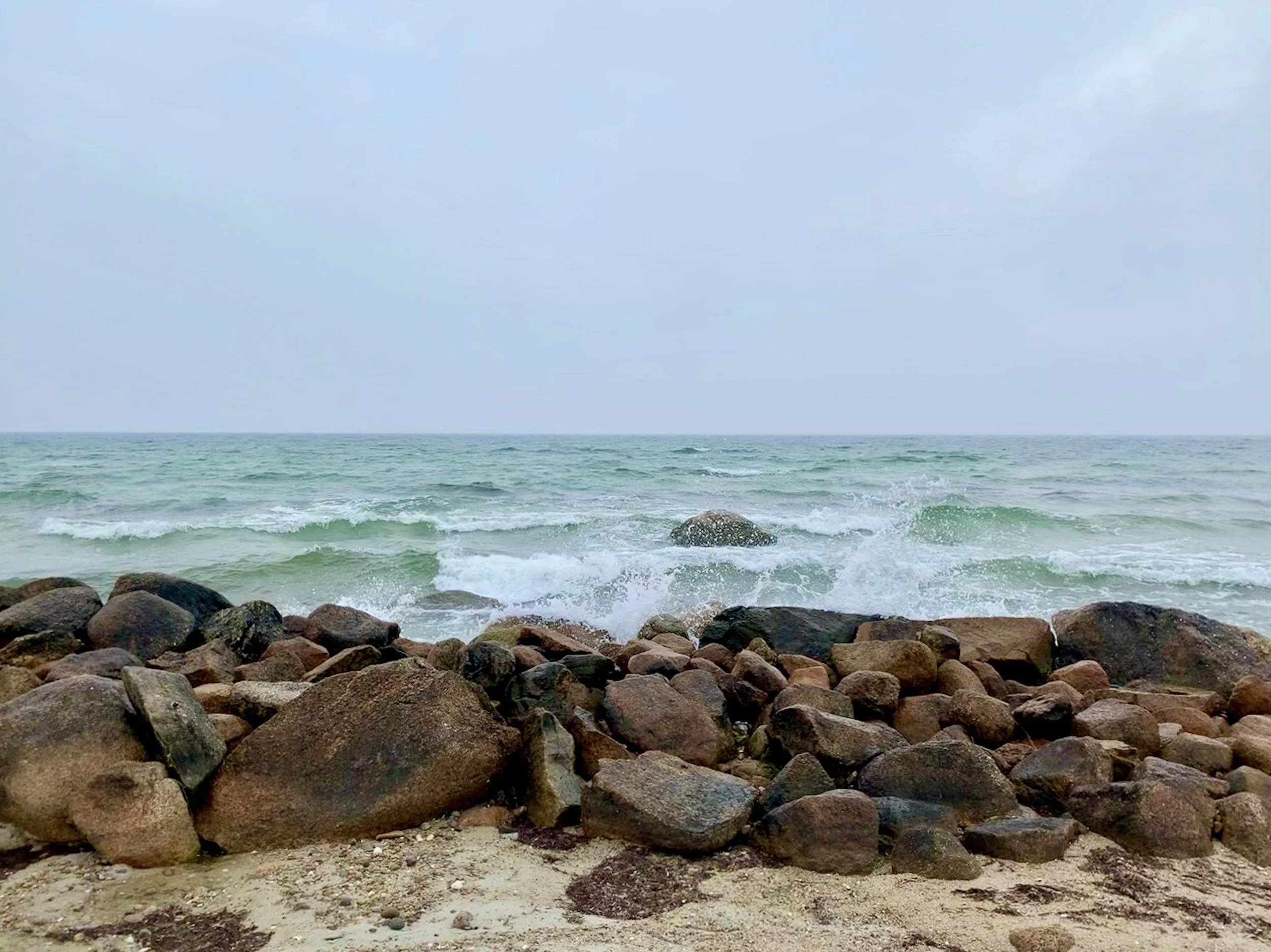 A rocky beach with large stones at the shore, ocean waves, and a cloudy sky.