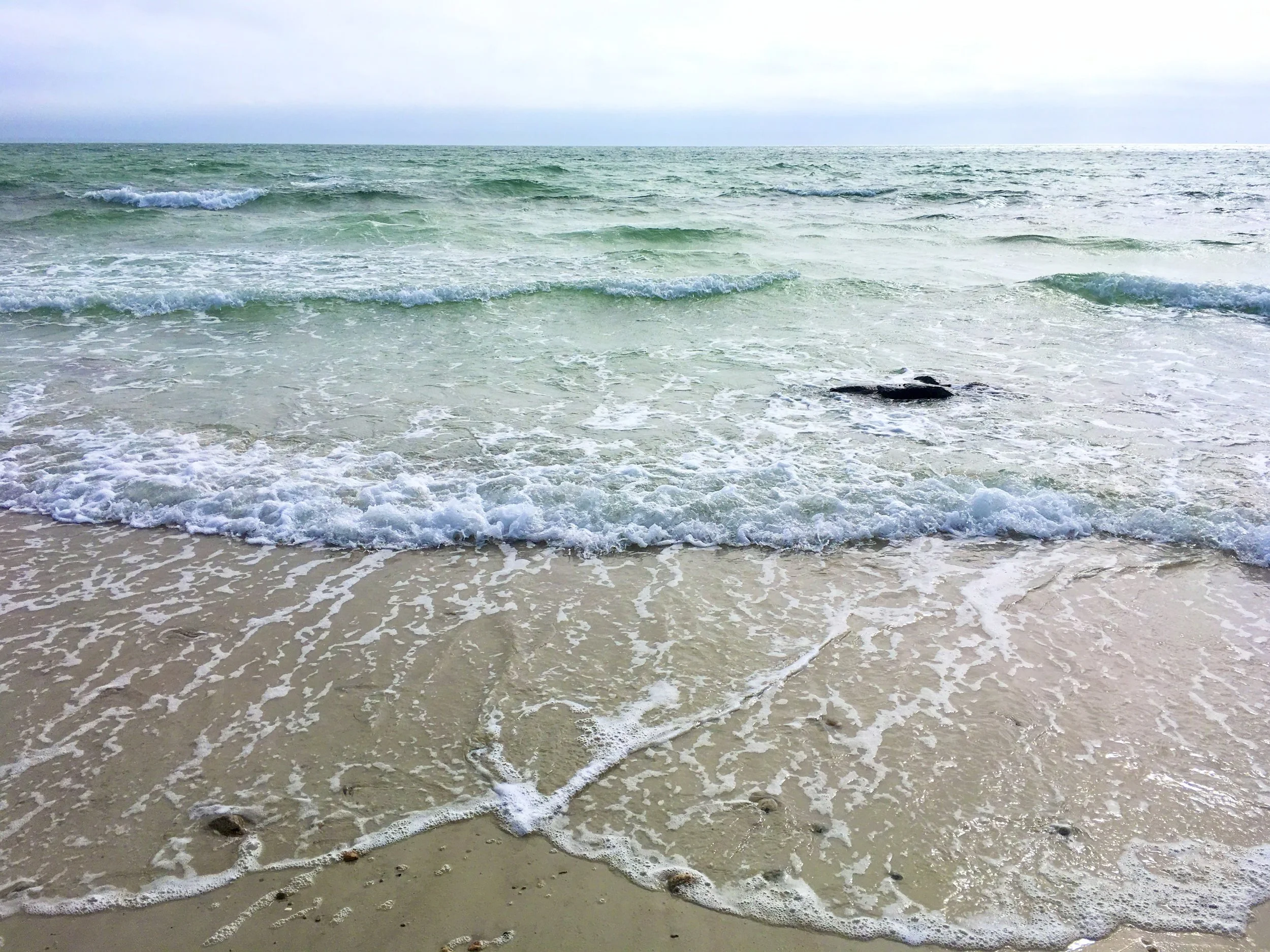 Waves crashing on a sandy beach with rocks and shells, under a cloudy sky.