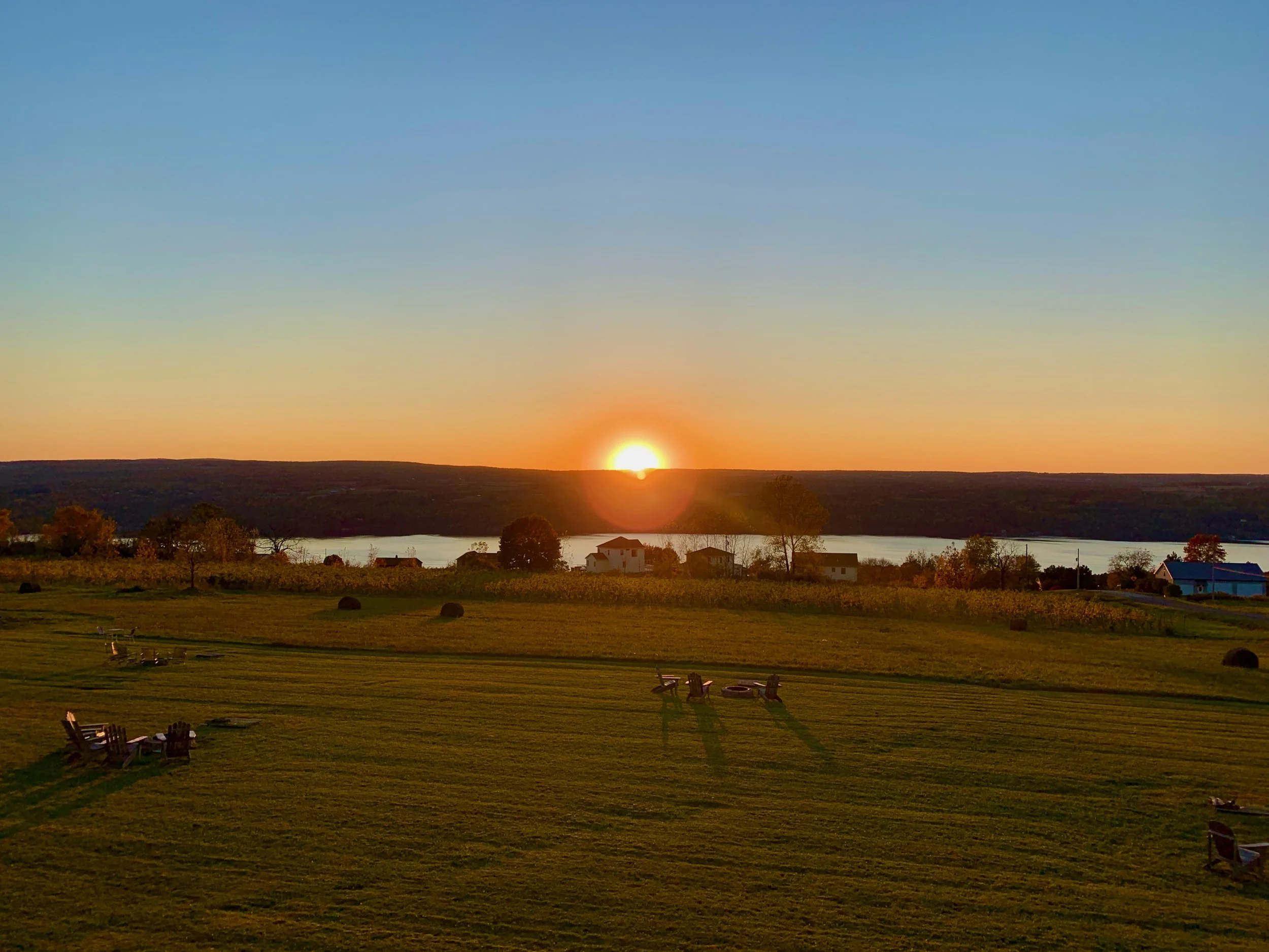 Sunset over a rural landscape with a lake, scattered trees, houses, and benches in the foreground.