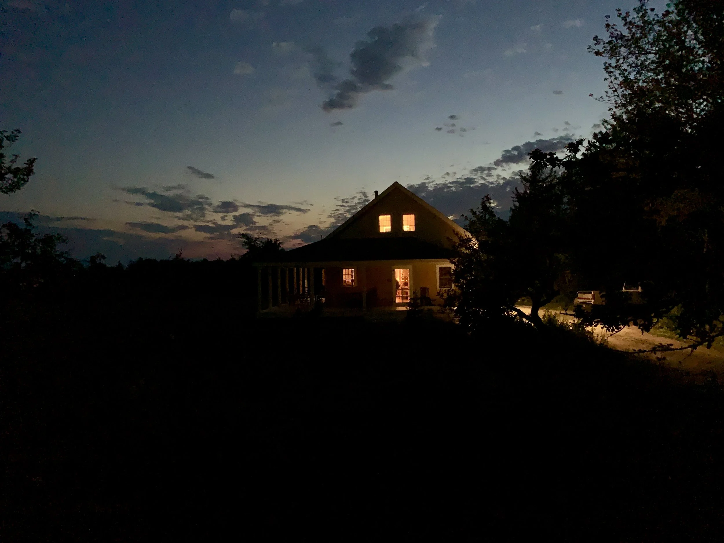 A house with two illuminated windows at night, surrounded by trees and a partly cloudy sky.