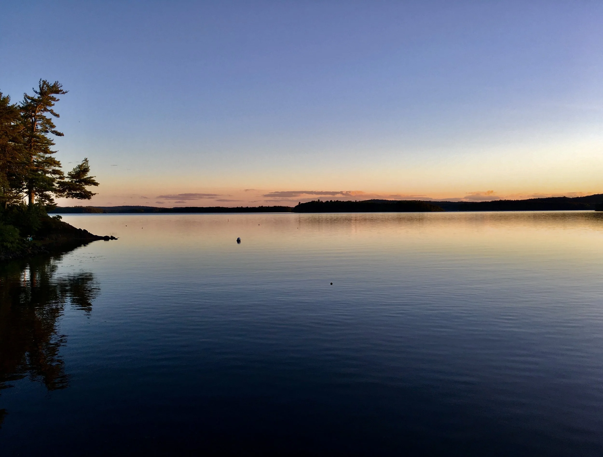 A tranquil lake during sunset with a clear gradient sky and calm water, bordered by trees on the left and distant land on the horizon.