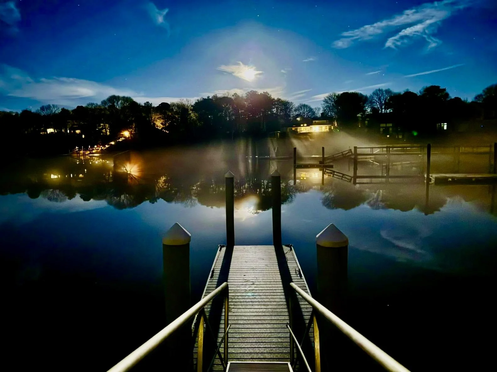 Nighttime view of a dock extending into calm water, with reflections of trees, houses, and the moon on the water's surface and a misty atmosphere.