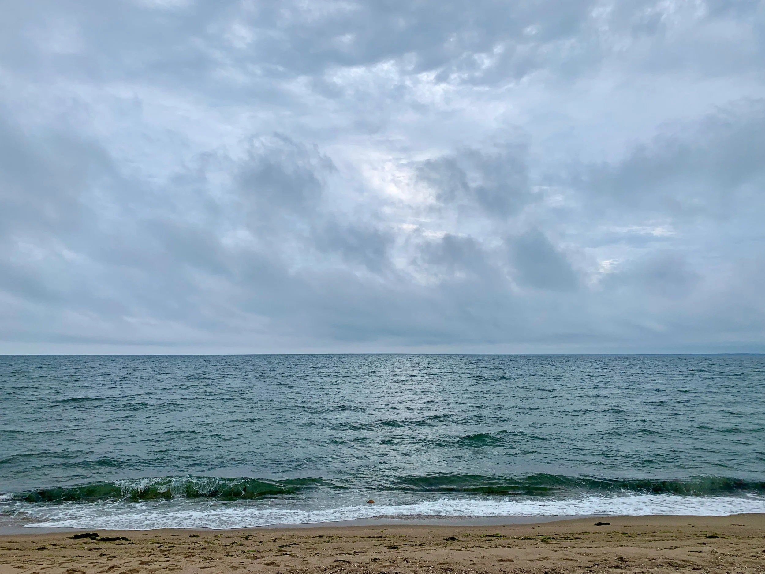 View of a cloudy sky over the ocean with gentle waves and a sandy beach in the foreground.