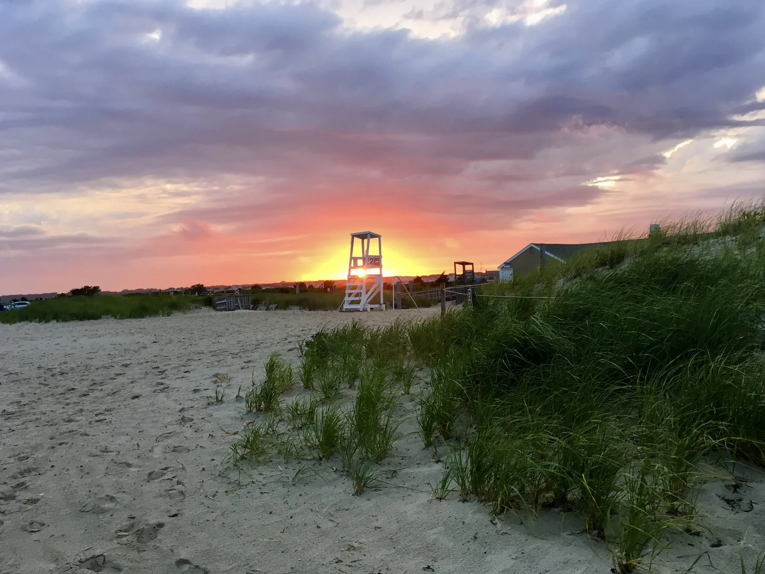 Sunset on a beach with a lifeguard chair, grassy dunes, a building, and cloudy sky.