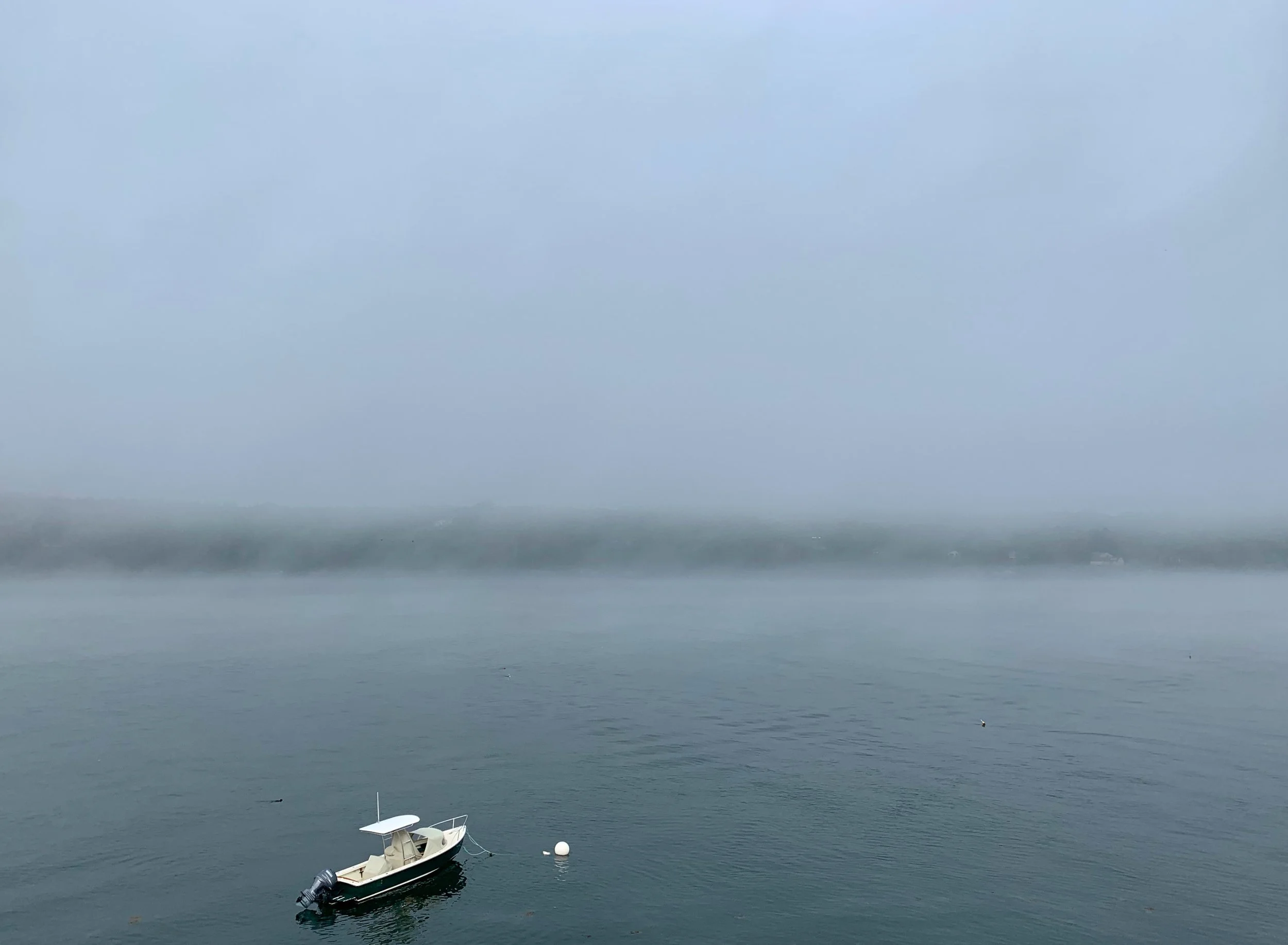 A small boat with an outboard motor floating on calm water near a white buoy, with misty or foggy weather and a distant shoreline.