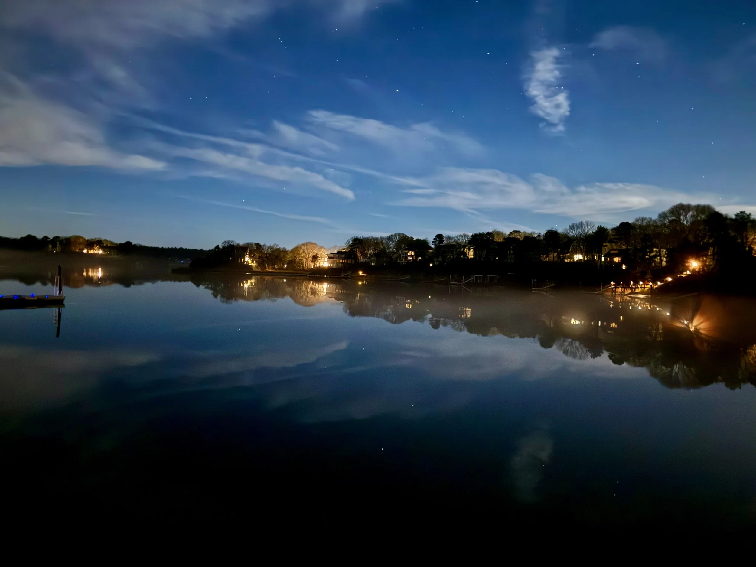 Nighttime scene of a calm body of water reflecting houses with lit windows along the shoreline, a boat docked on the left, and a sky filled with stars and wispy clouds.