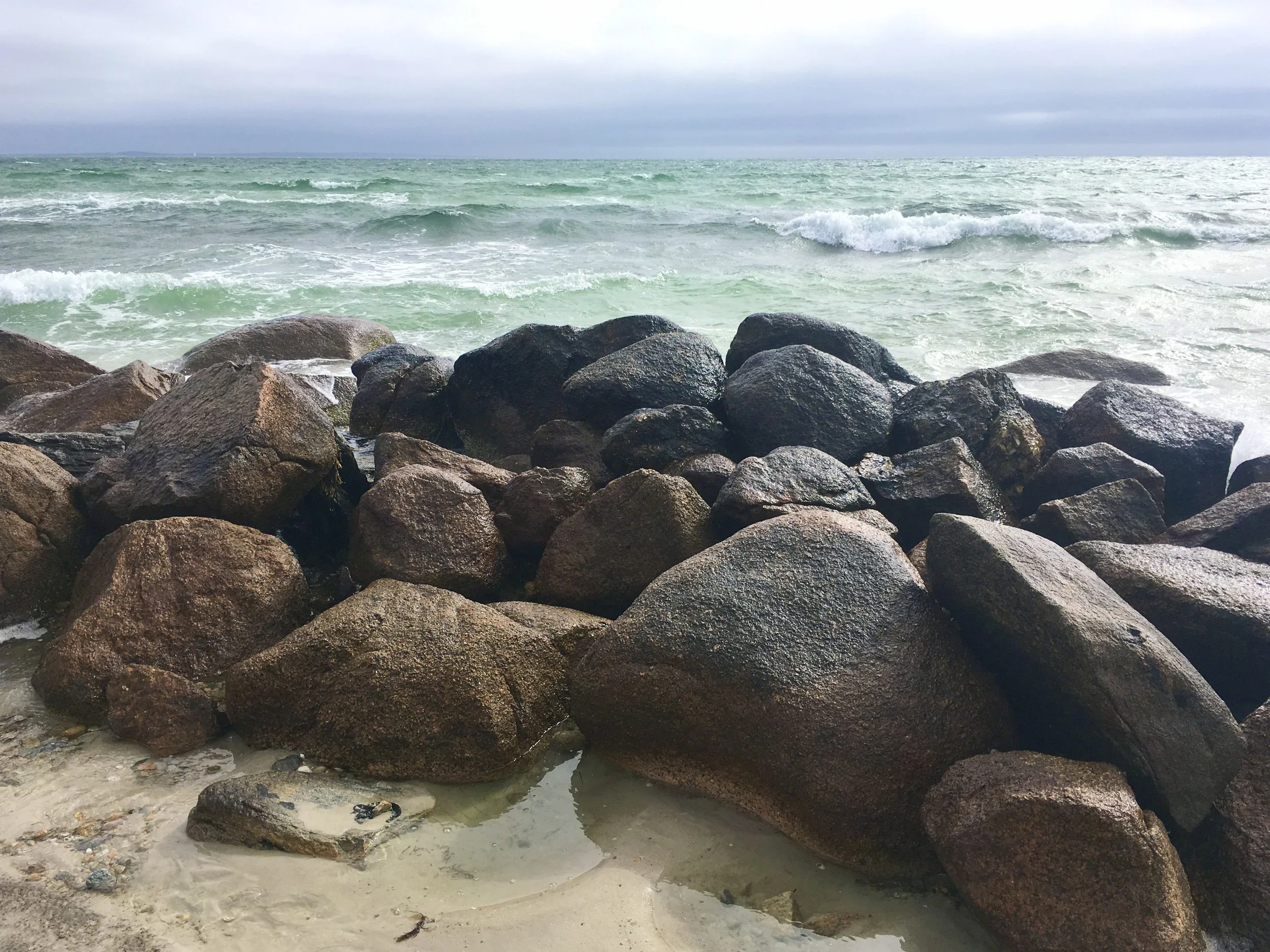 A rocky shoreline with large dark rocks in the foreground, small pools of water, and waves crashing against the rocks on a cloudy day at the ocean.