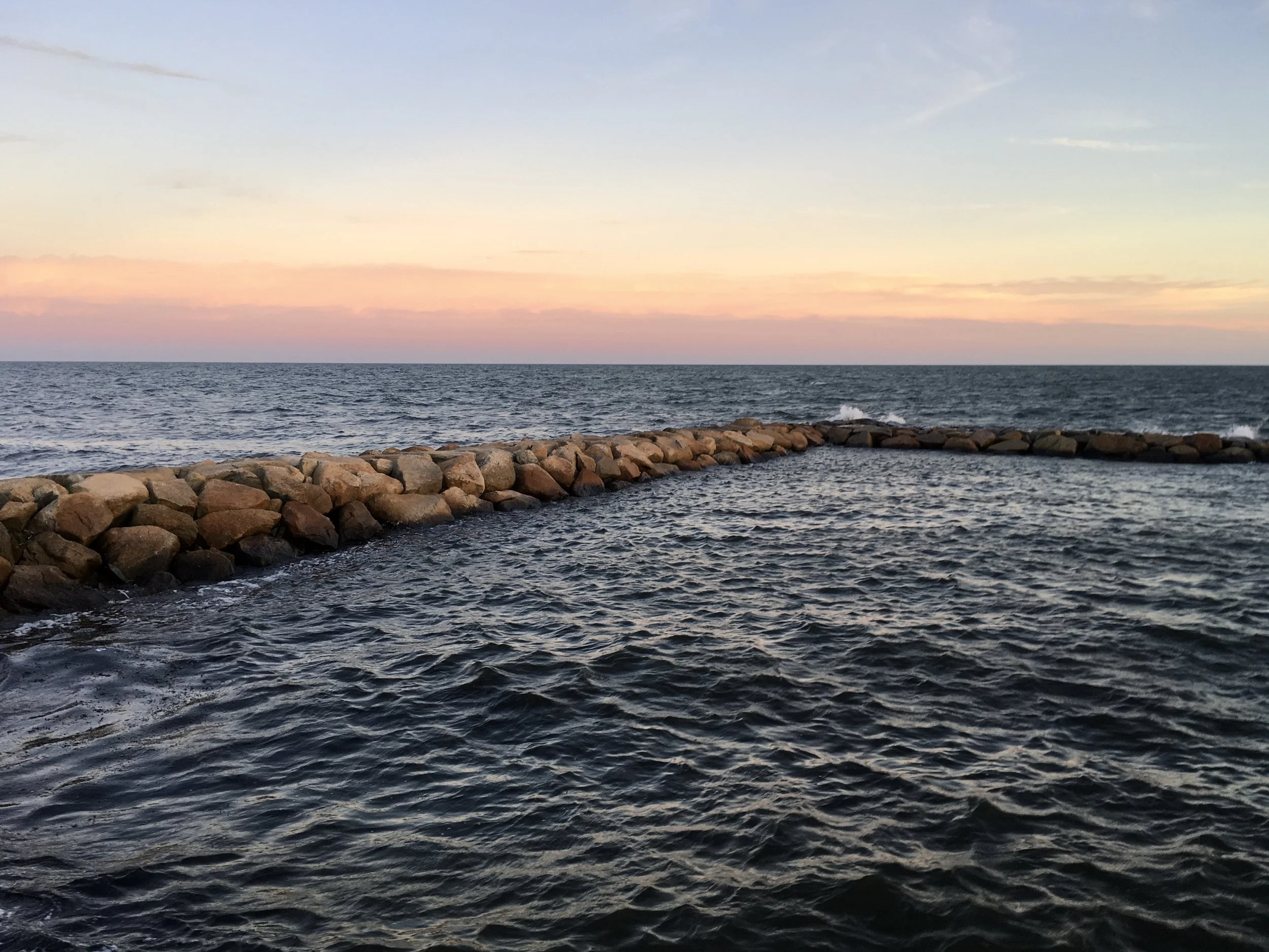 Calm ocean water with a rocky breakwater extending into the sea, under a sky with soft pastel colors from a sunset or sunrise.