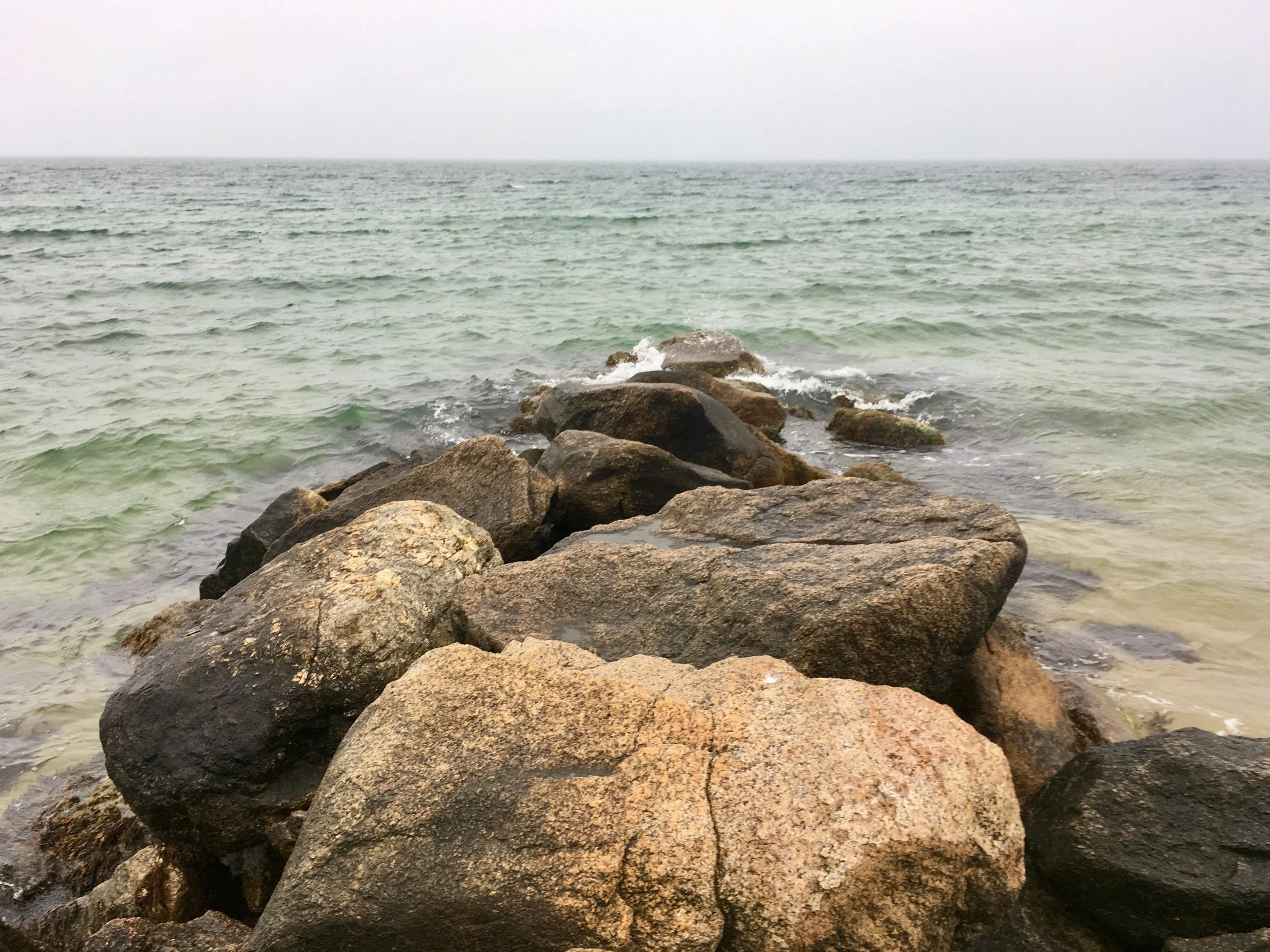Rocky shoreline with large boulders in the foreground and the ocean extending to the horizon under an overcast sky.