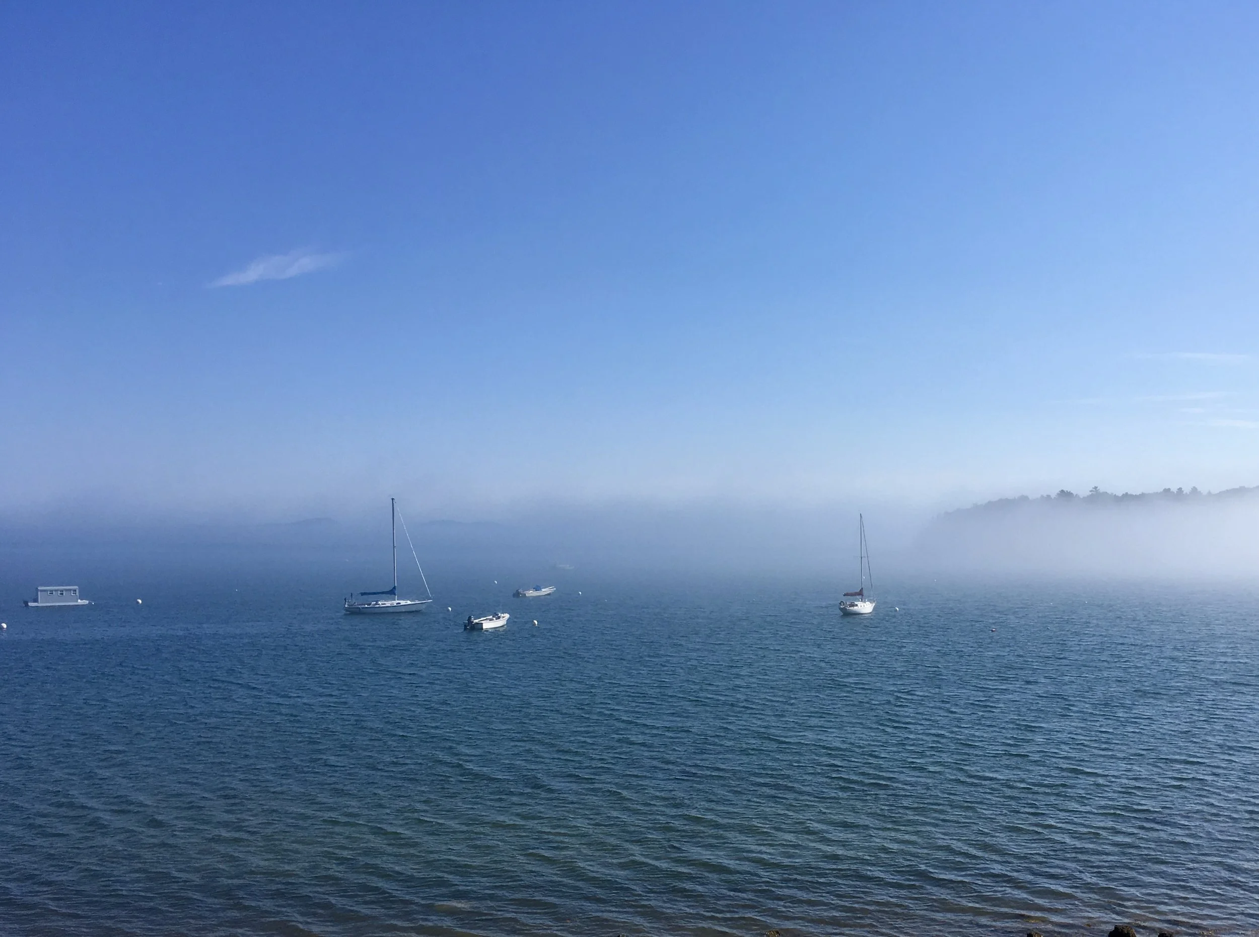 Calm ocean with several sailboats anchored, distant foggy land in the background, and a bright blue sky above.