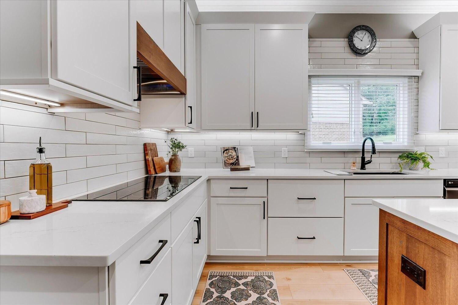 Modern white kitchen with black hardware, a dark faucet, a window with blinds, wall clock, and wooden accents.