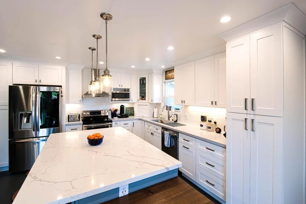 Modern white kitchen with marble island, stainless steel appliances, pendant lights, and under-cabinet lighting.