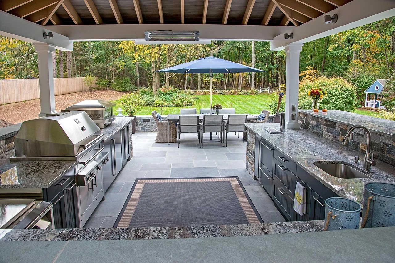 Outdoor kitchen and dining area with a view of a backyard garden, an umbrella, and patio furniture under a roof.
