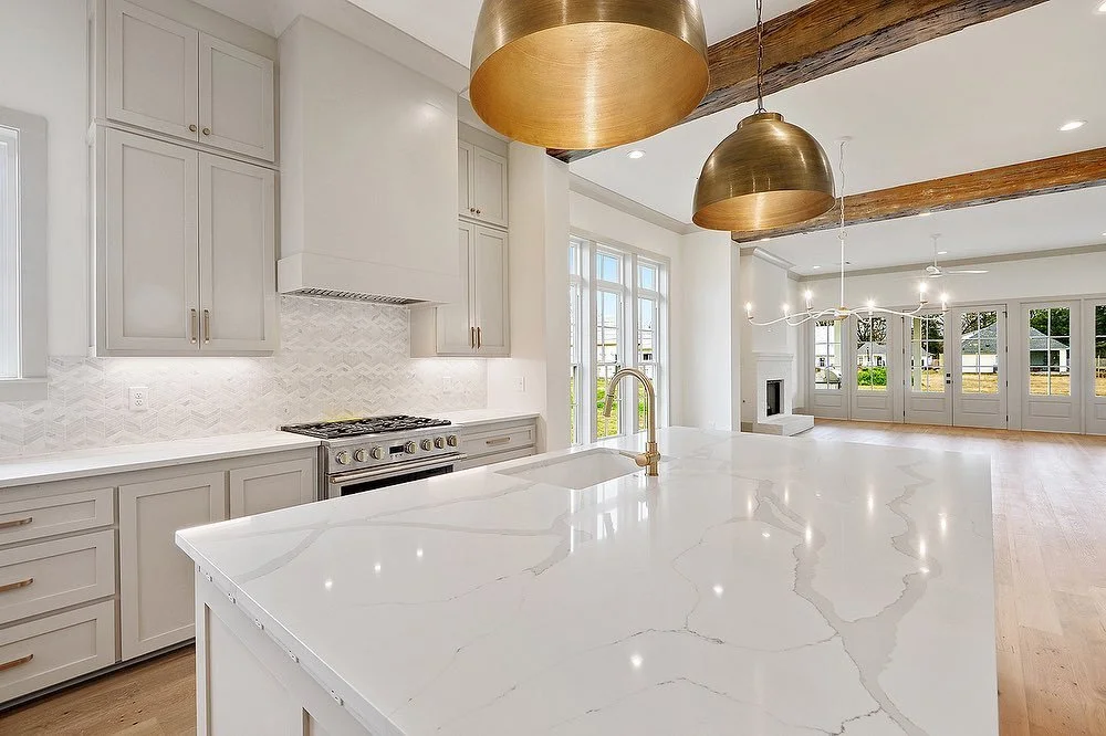 Modern kitchen with white cabinetry, marble island, and pendant lights, open to a living area with large windows and hardwood floors.