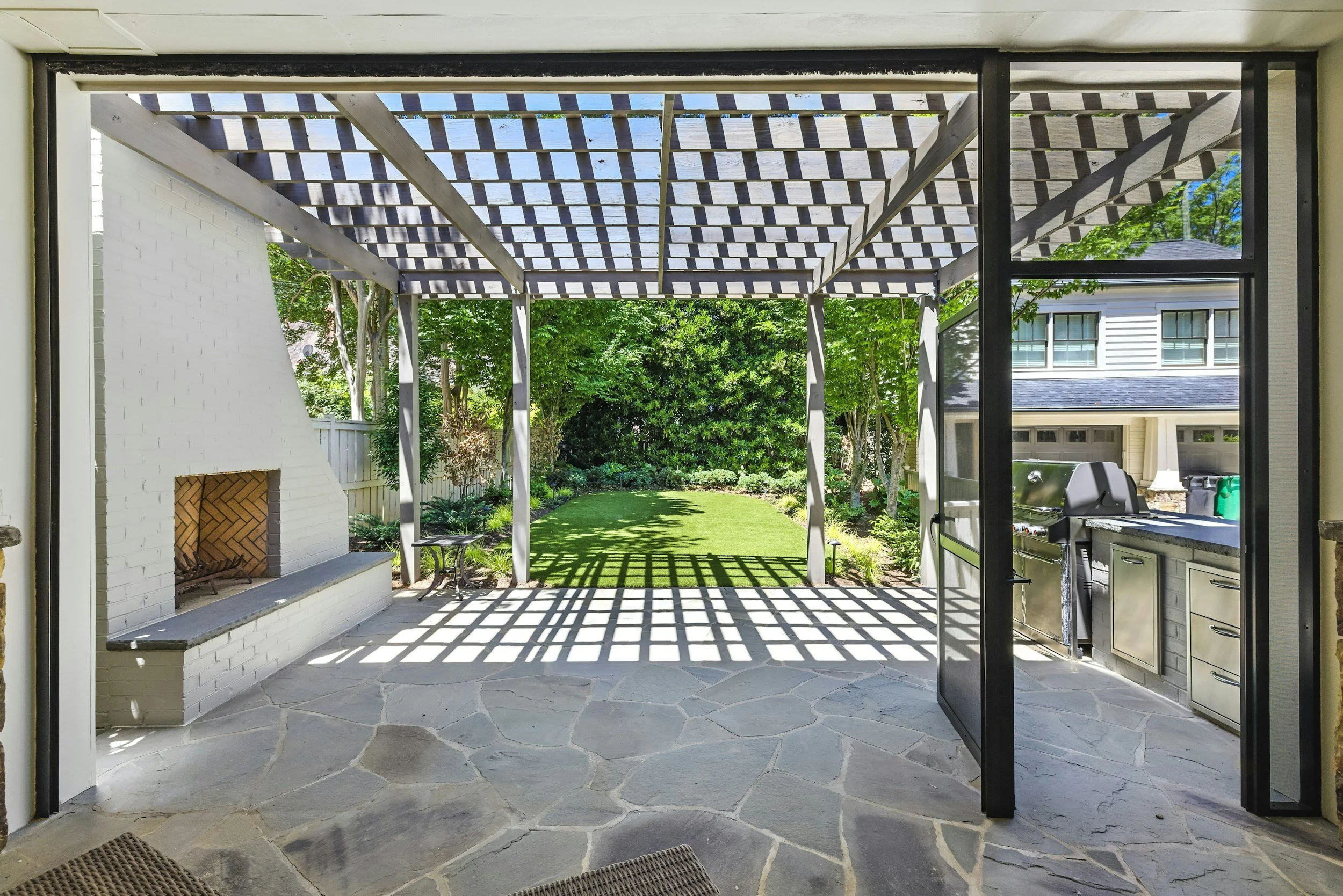 View of a backyard patio remodeling project in Frisco, Texas with a wooden pergola, green grass, trees, and a neighboring house in the background, seen from inside a screened porch area.