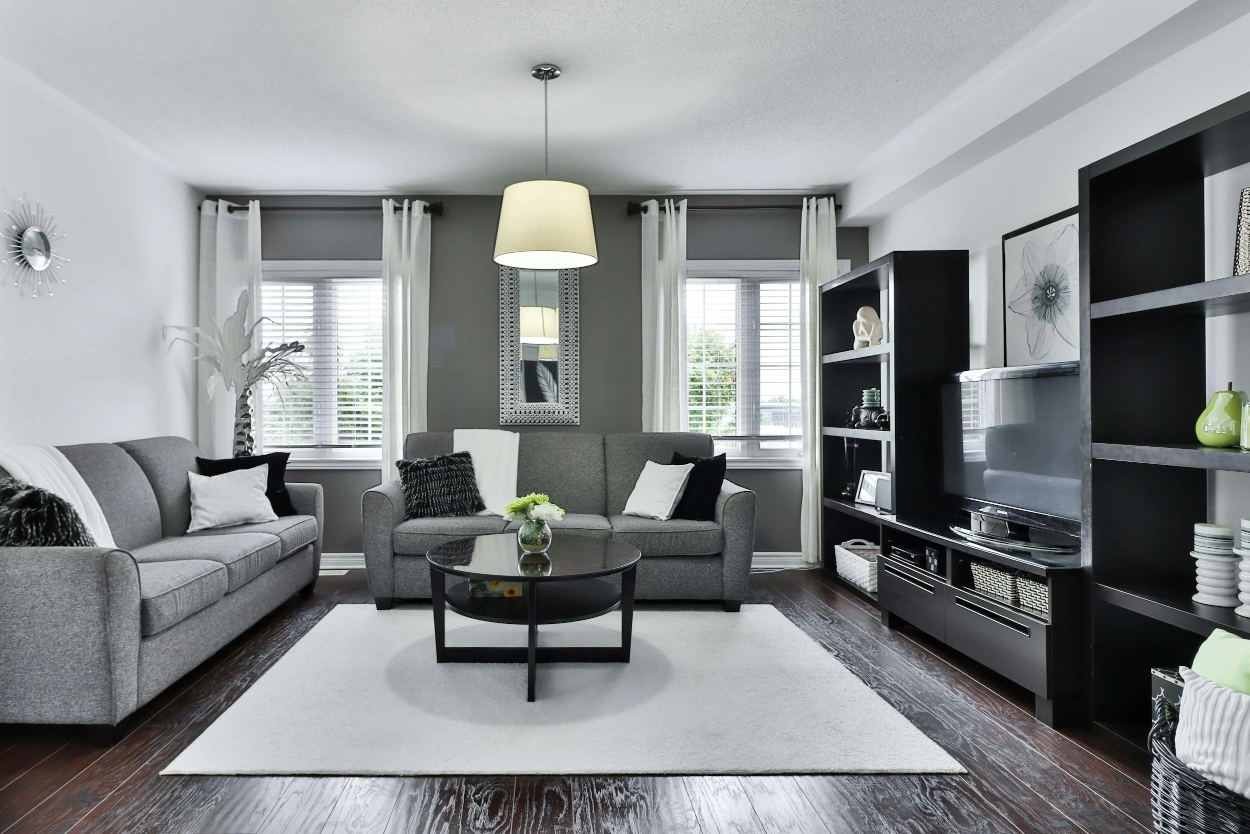 Living room with gray sofas, black coffee table with flower arrangement, white area rug, black entertainment center with TV, dark hardwood floors, white walls, large windows with white curtains, artwork, and decorative items.