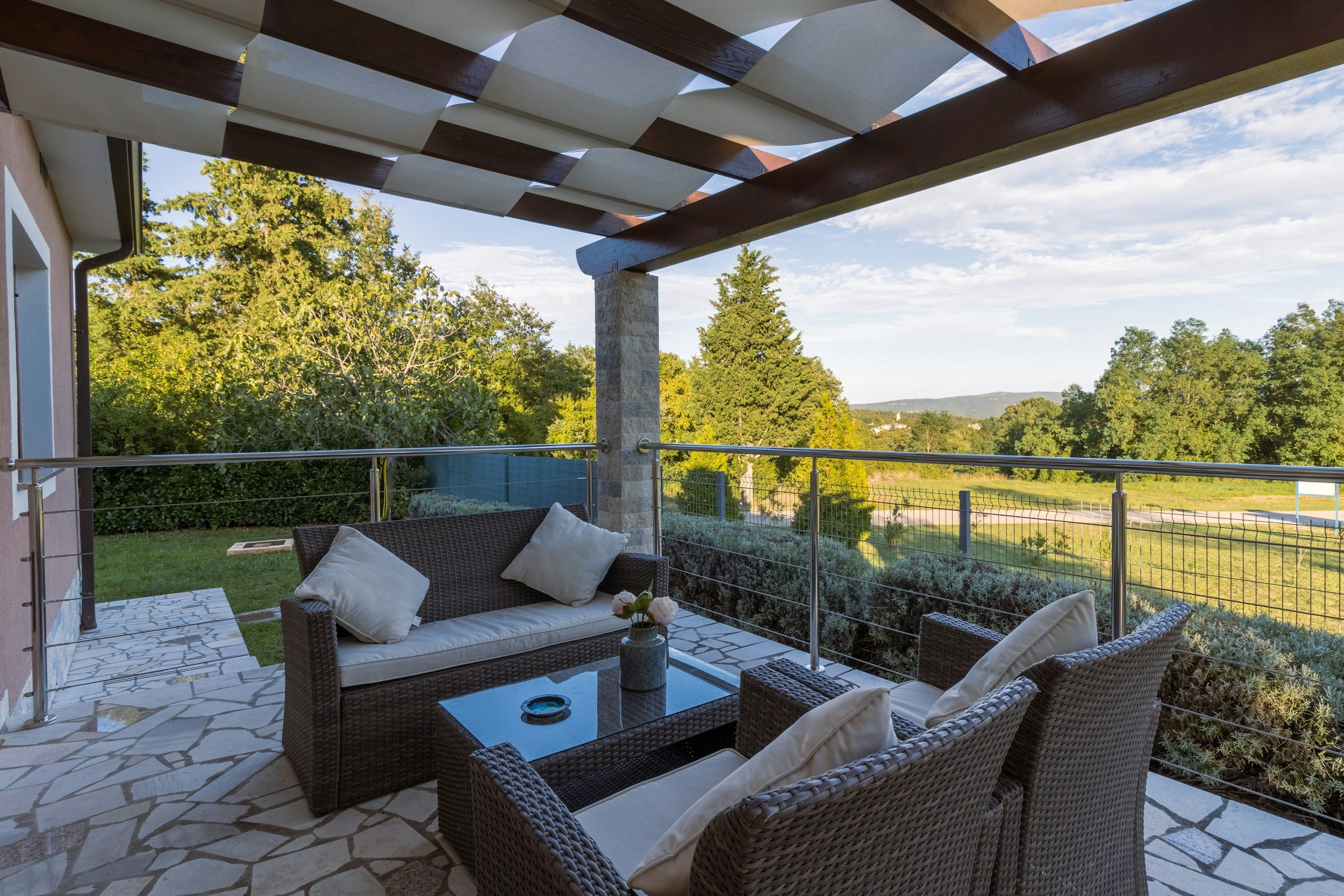 A covered outdoor patio with wicker furniture, cushions, and a glass-top coffee table. There is a view of trees and a distant landscape beyond a metal railing.