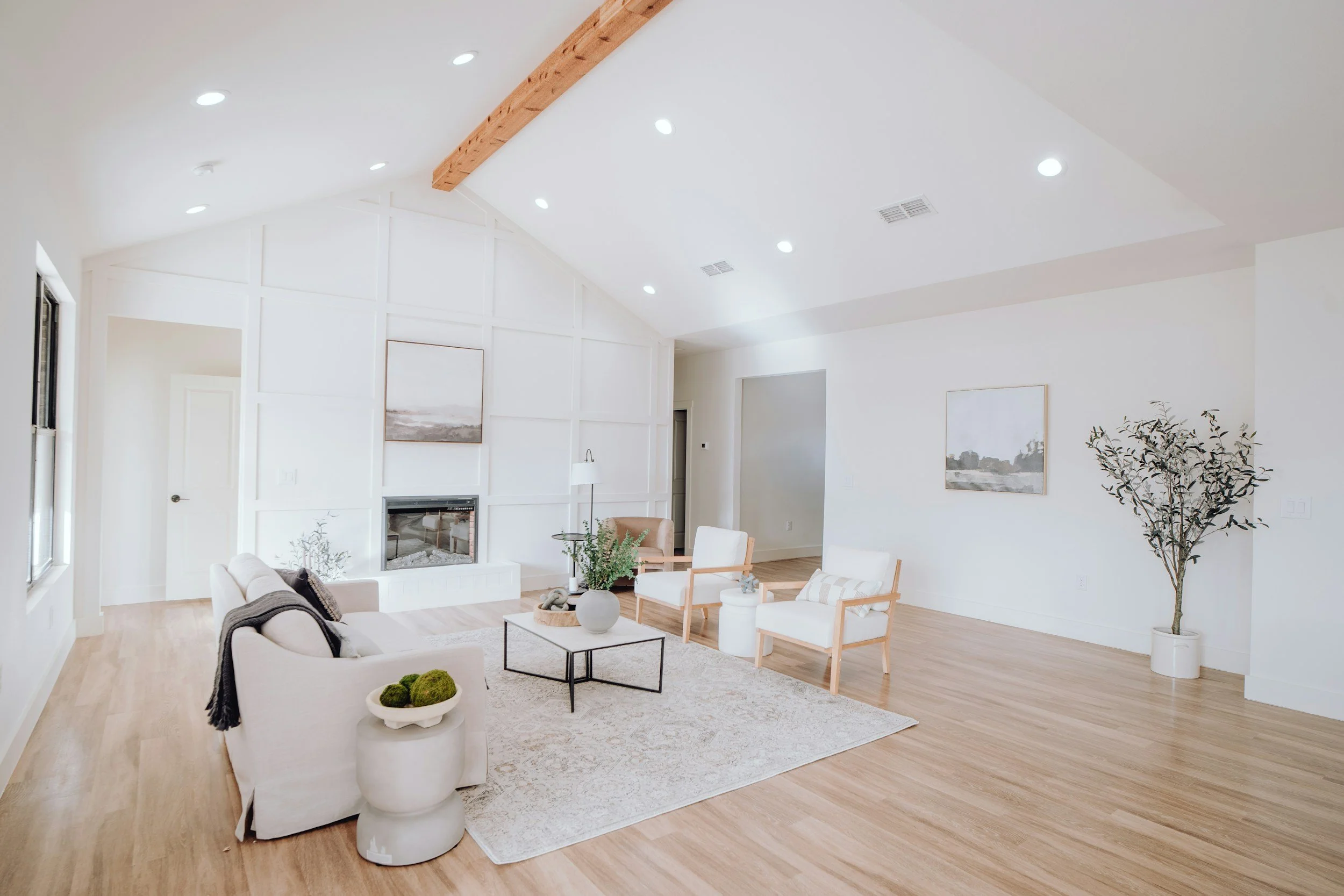A spacious living room with white walls, light wood flooring, and a vaulted ceiling with recessed lighting and a wooden beam. The space features a cream-colored sofa, white armchairs with wooden frames, a black coffee table, and decorative plants and