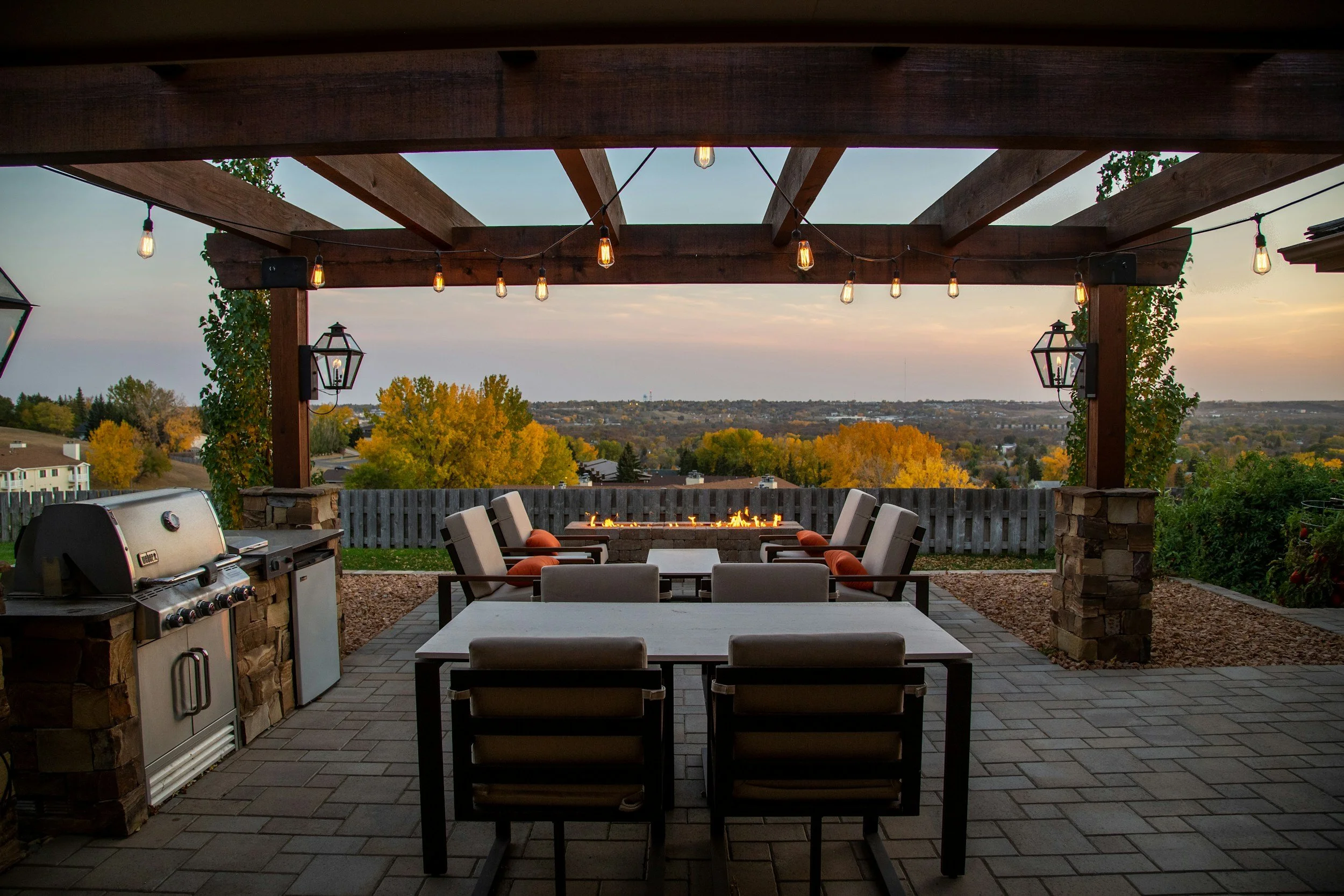 Outdoor patio with seating area under a wooden pergola decorated with string lights, a fire pit in the background, and a barbecue grill on the left, overlooking a scenic view of trees and hills at sunset.
