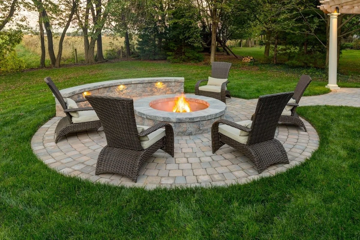 Outdoor patio remodeling done in Plano, Texas with a circular stone fire pit surrounded by five wicker chairs with cushions, set on a paved stone surface in a backyard with green grass and trees.