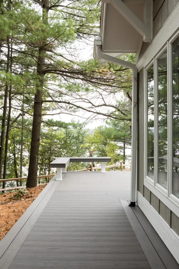 View of a wooden deck attached to a house, with trees in the background and part of the house's exterior wall and window visible.