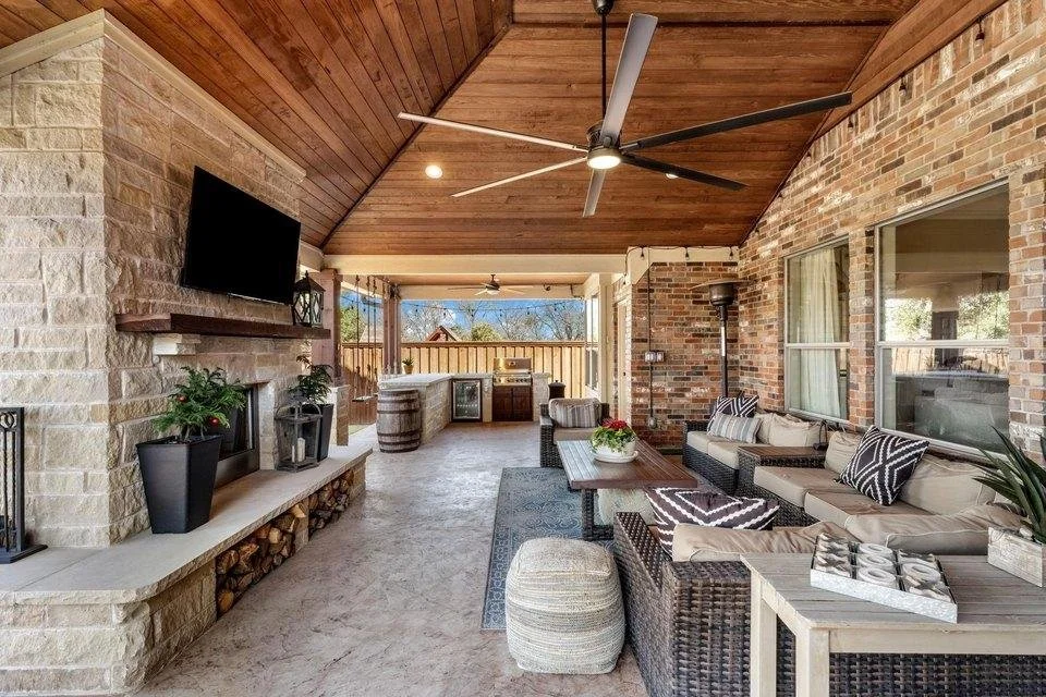 Covered porch with a cozy seating area including a sectional sofa with throw pillows, a coffee table with a flower arrangement, and a beige pouf. There's a wall-mounted TV above a fireplace with decorative logs and plants. The porch has a wood-paneled ceiling with a ceiling fan, brick walls, and sliding glass doors leading to the interior of the house. The background shows a fenced yard and outdoor scenery.