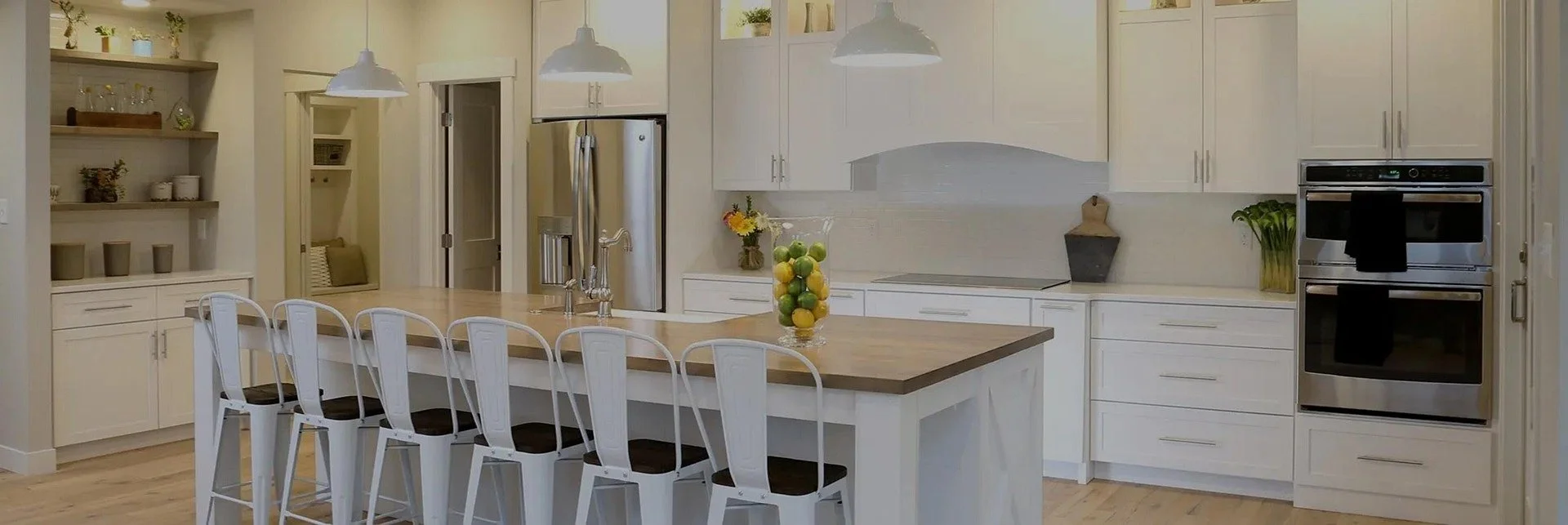 Bright modern kitchen with white cabinets, wooden countertops, stainless steel refrigerator, built-in oven, and a kitchen island with bar stools. Decor includes a bowl of fruits, flowers, and potted plants.